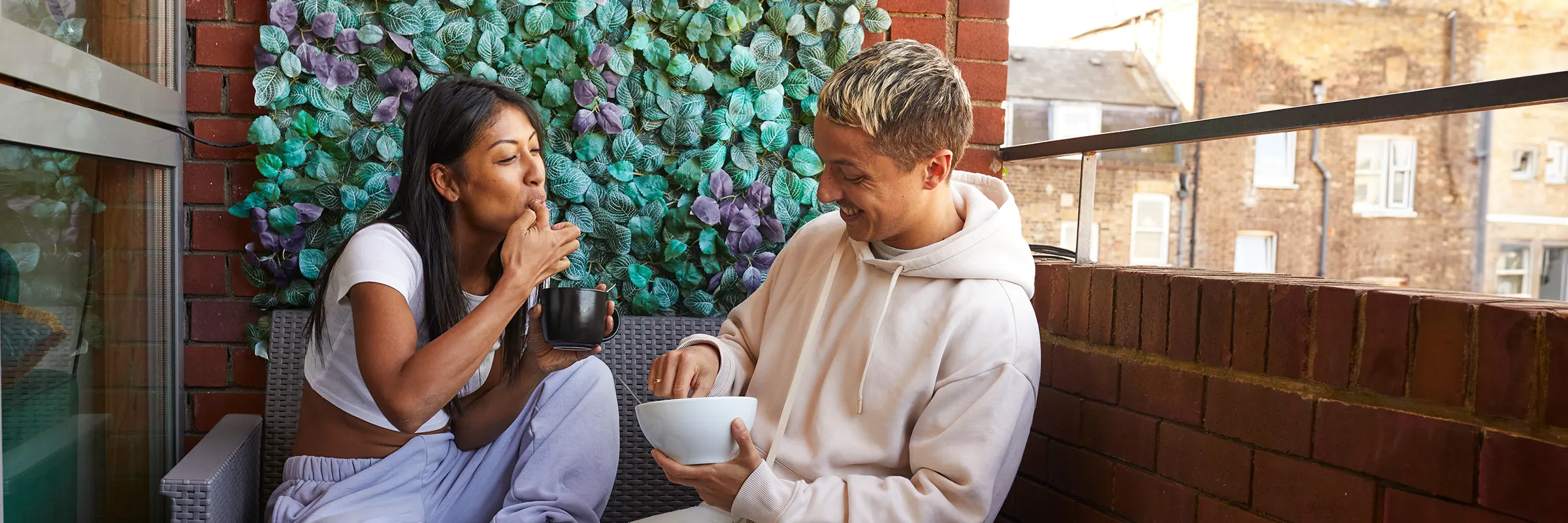 A young woman and man sit next to each other on a small balcony enjoying breakfast.