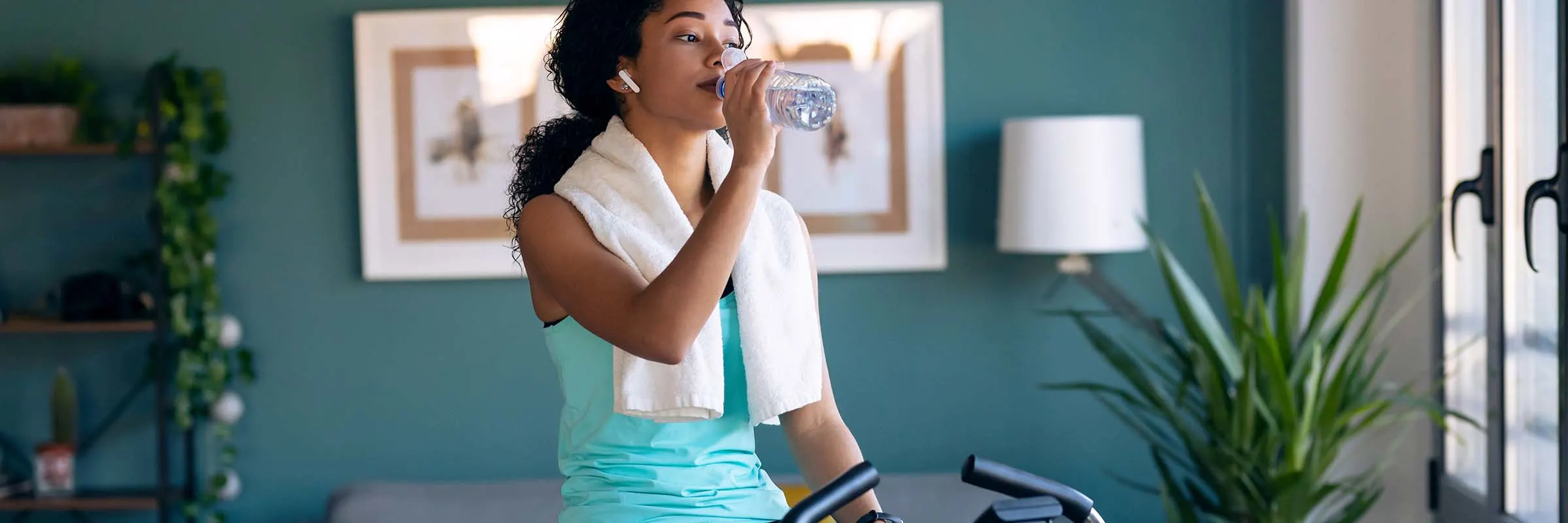 Young woman is drinking out of a water bottle while sitting on a stationary bike in her home.