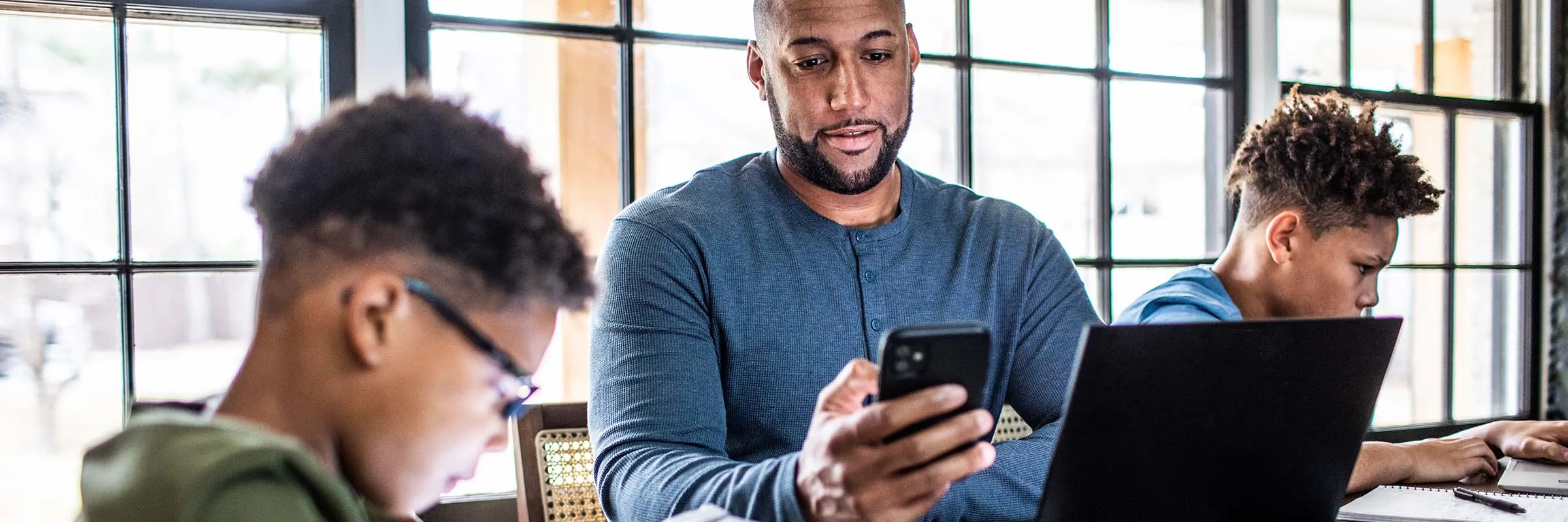 Father and two young sons sitting at a table with their tech. Son on the left has a tablet and son on the right has a laptop. The father is looking at his smartphone with his laptop open in front of him.