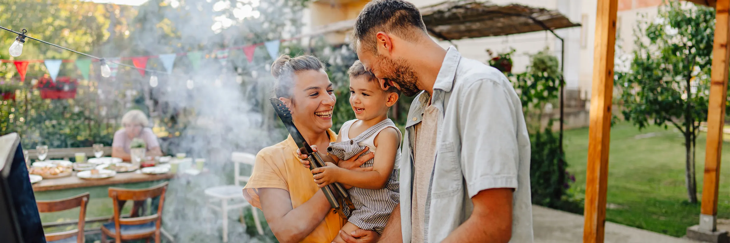 Family of three using an outdoor grill, the mother holds a child and the father looks at them while smiling.  