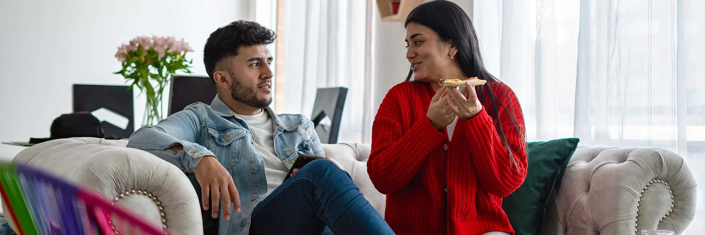 A young man and woman sit on a couch in their living room and eat pizza.