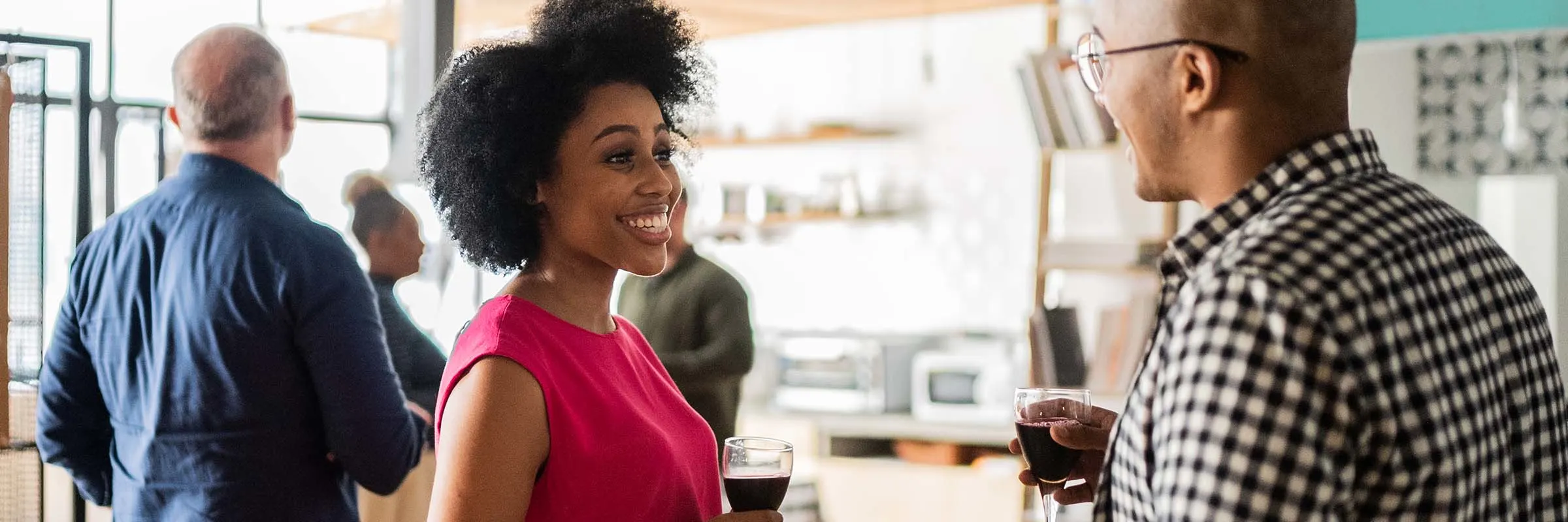 A woman is smiling and holding a glass of wine while speaking with a coworker in an office.