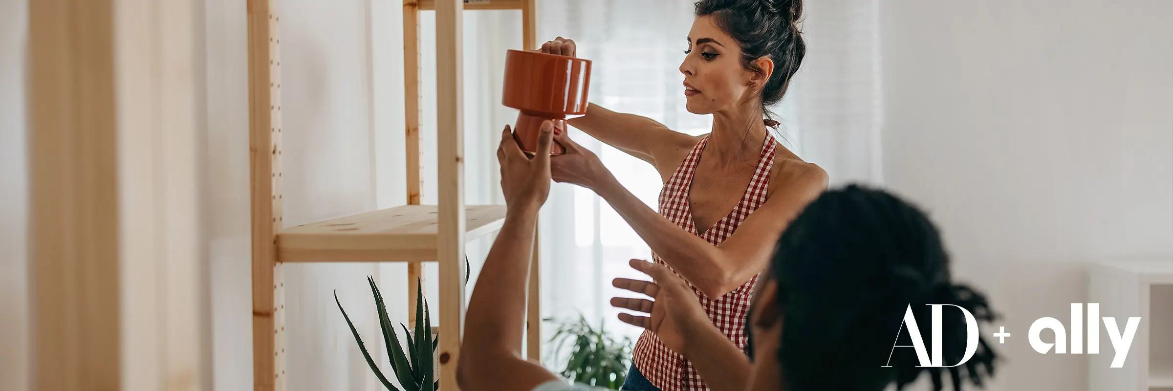 A young man hands a woman a vase to set on the top level of the bookshelf. 