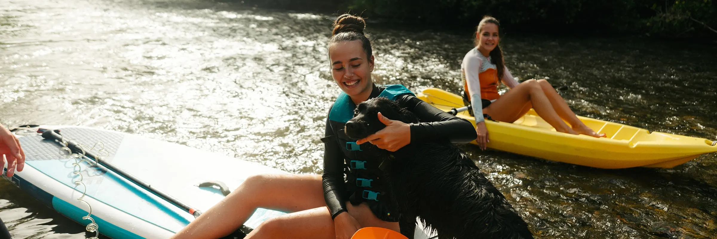 A young woman, surrounded by two friends, sits on a paddle board in shallow water while petting a dog. 