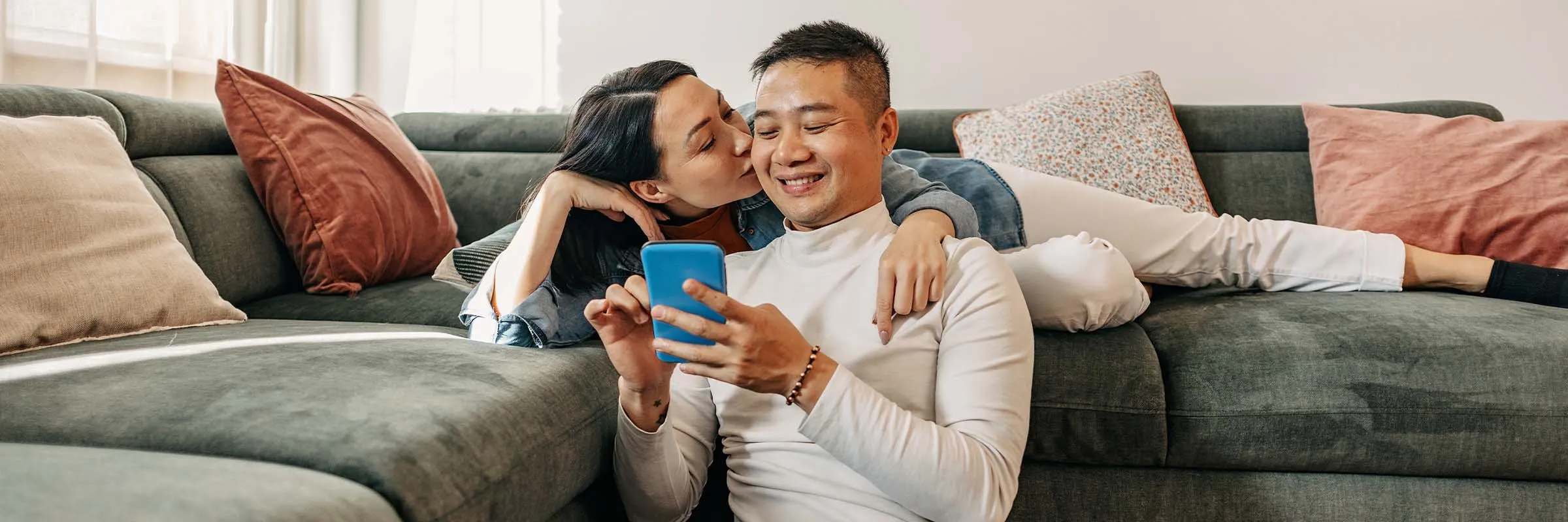 Photo of a woman kissing her boyfriend, lying on the sofa, while he shows her something on the phone