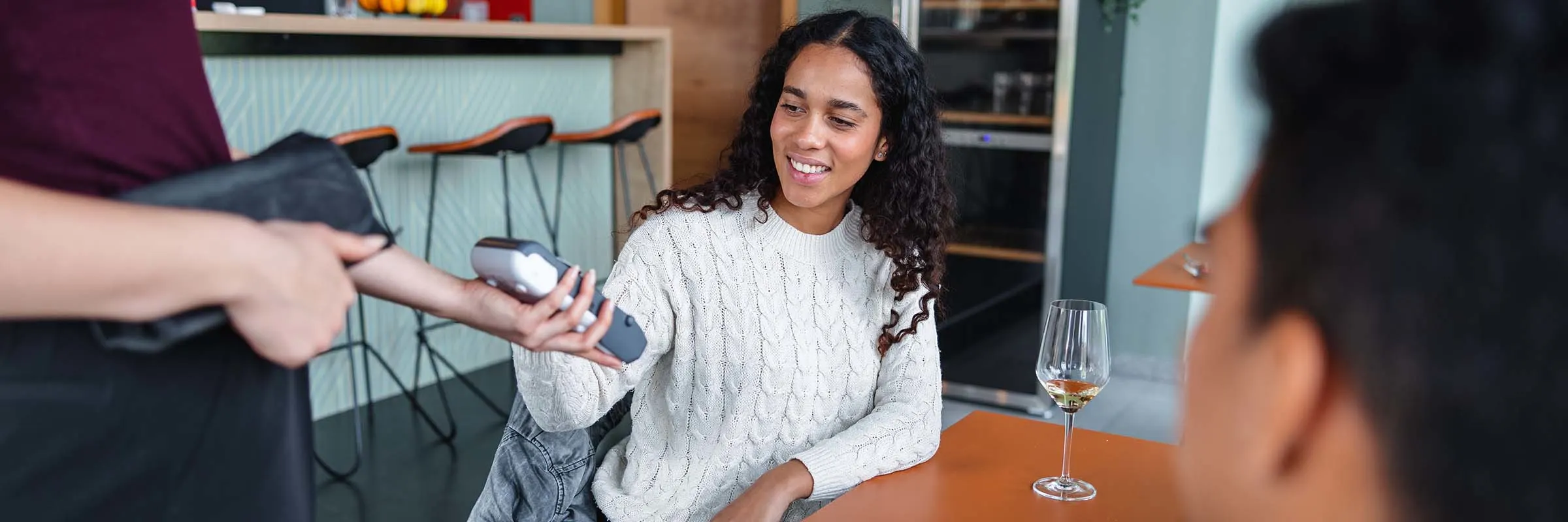 A woman pays for a glass of wine at a restaurant using a card reader.