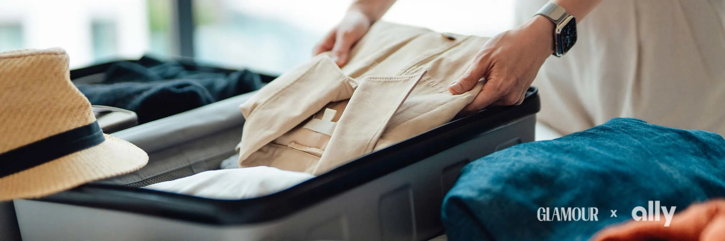 A person wearing a watch is packing a suitcase. There are clothes, a hat and a camera near the suitcase.