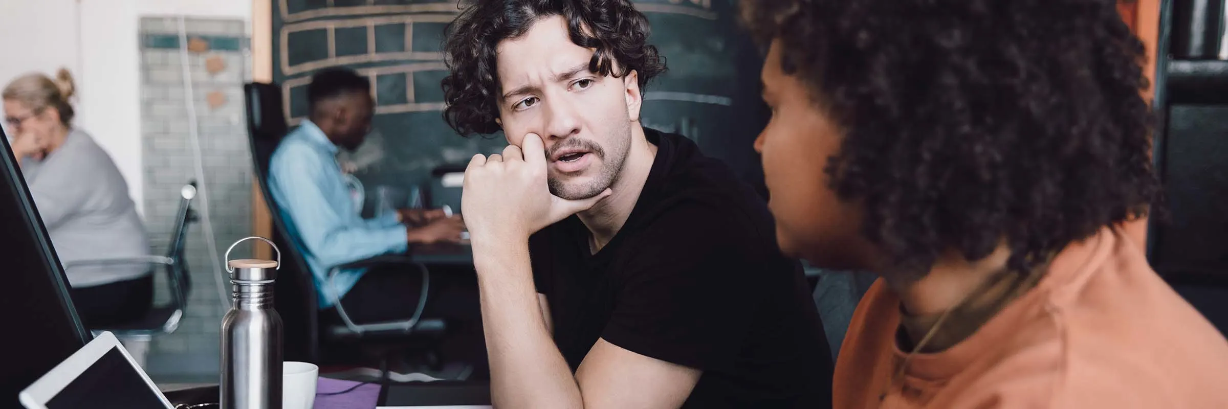 Two colleagues are having a conversation while sitting at a desk in front of a laptop.