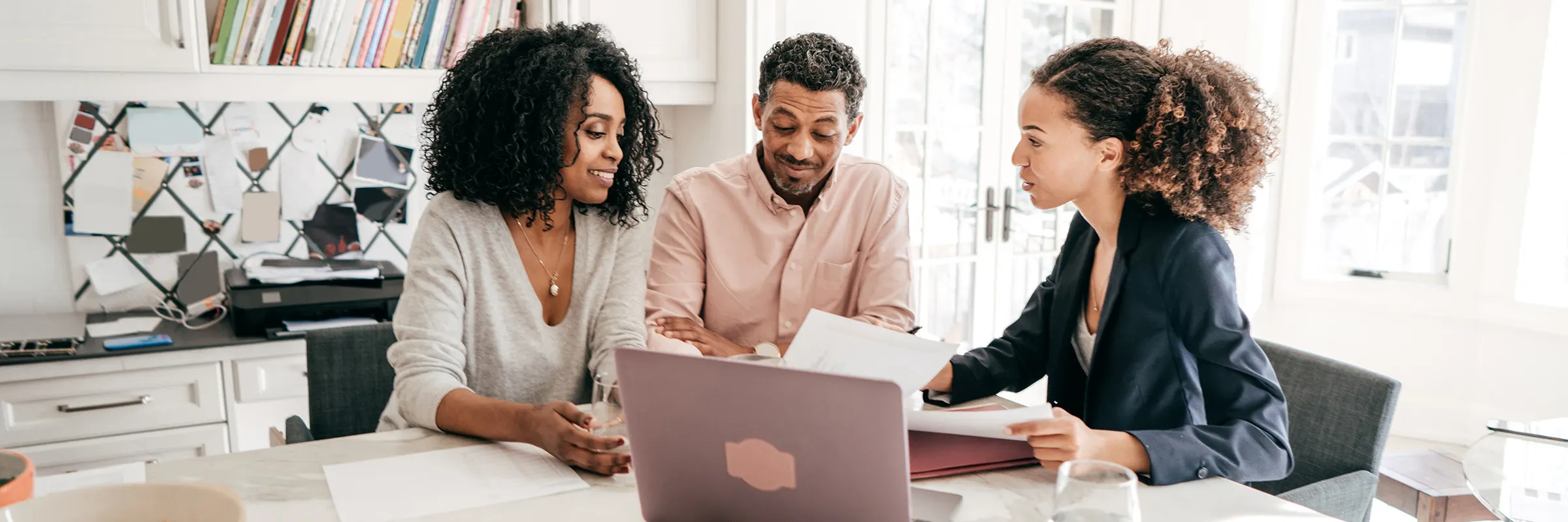 A young man and woman sit at a kitchen table with an advisor and laptop looking at paperwork.
 