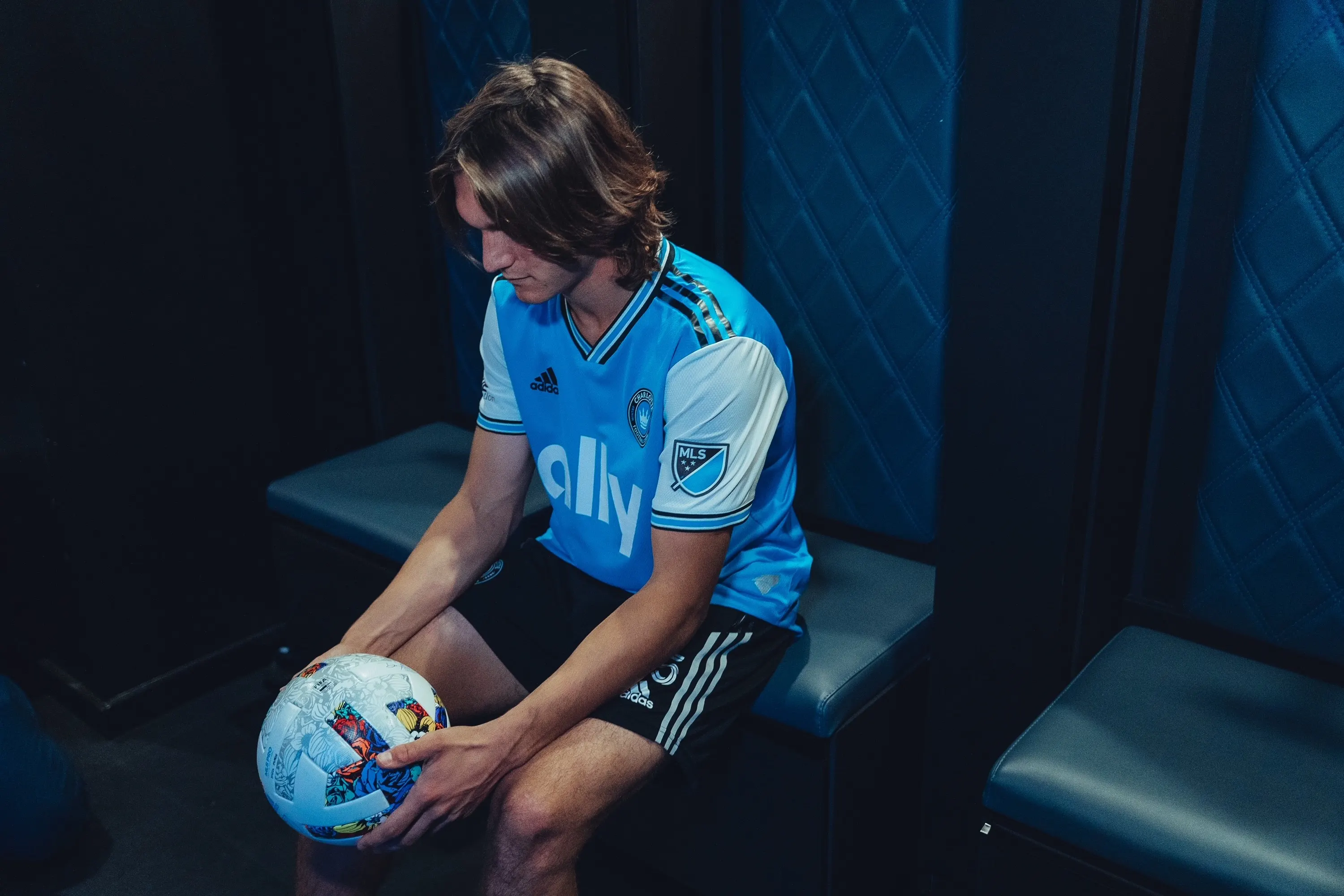 Soccer player Ben Bender sits in a locker room stall holding and looking down at a soccer ball. 