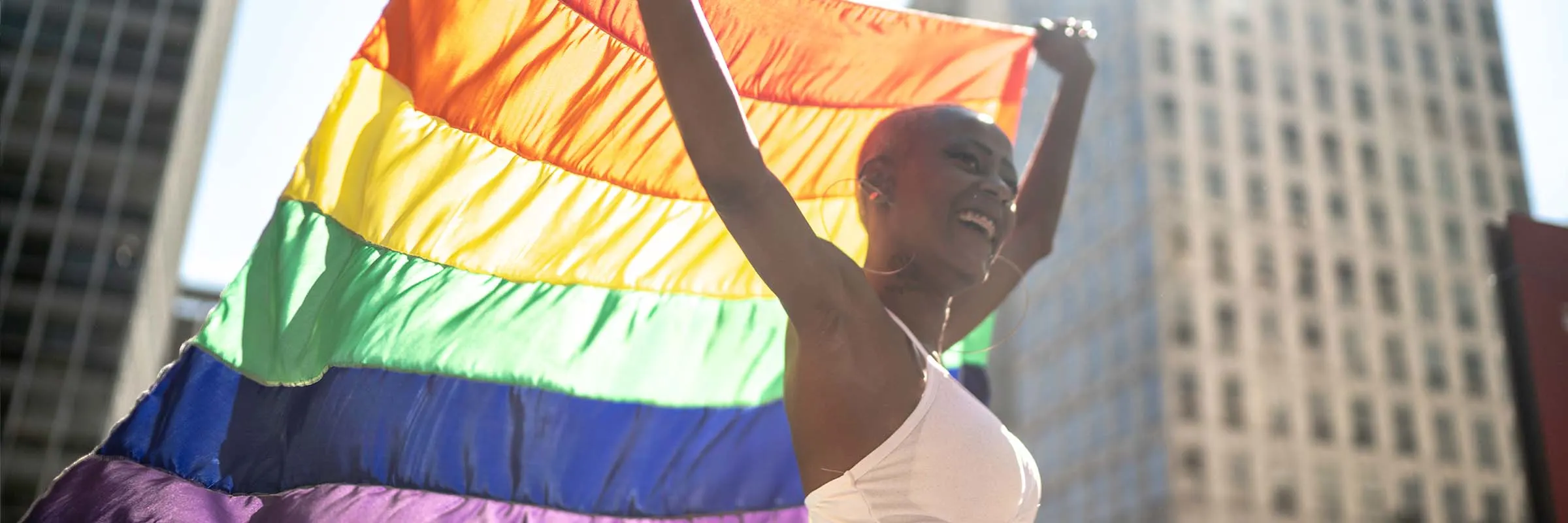 A woman is smiling and holding a rainbow flag above her head while walking in a city.