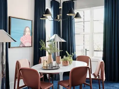 A chandelier hangs above a round dining room table that seats six. Long curtains frame two sunny windows.