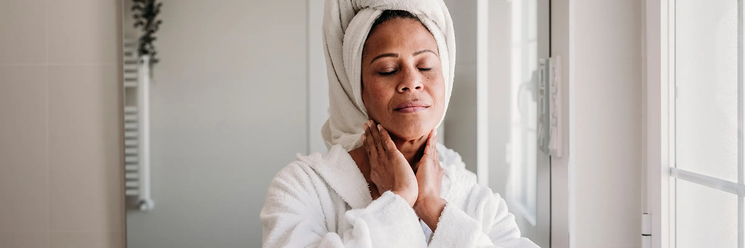 A woman rubbing her neck while wearing a robe with her hair wrapped in a towel.