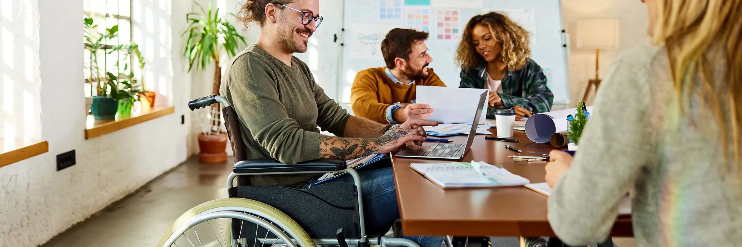 Happy creative man in wheelchair working on a computer while having a meeting with his colleagues at casual office.