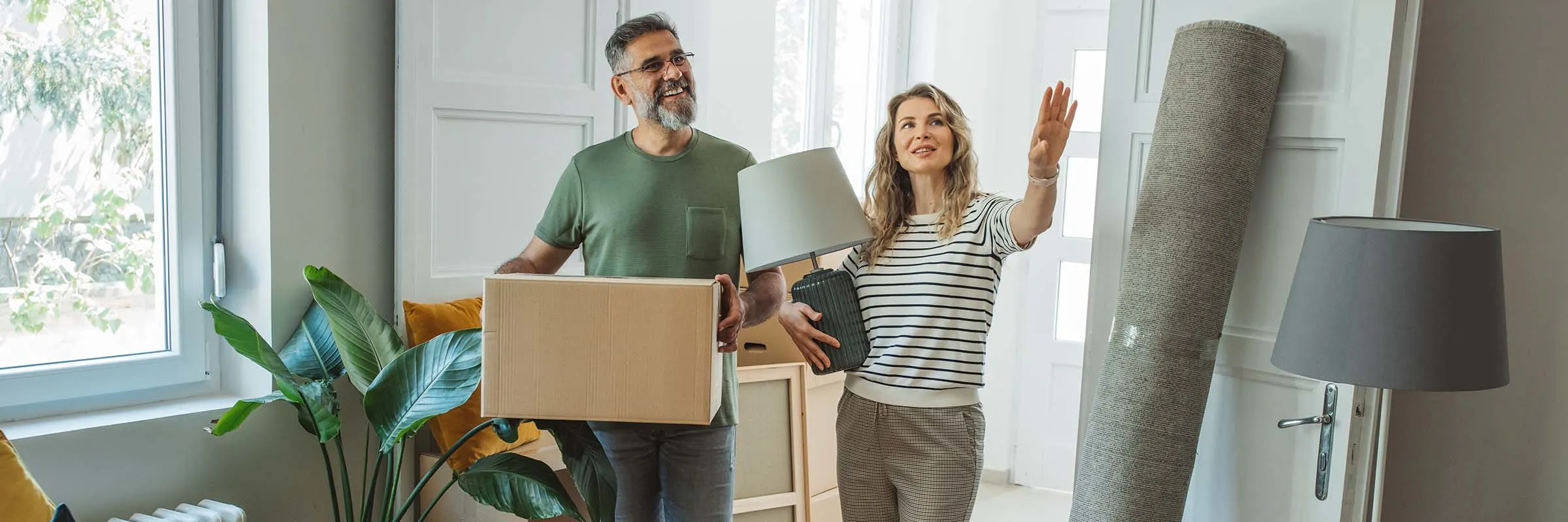 A man and woman are carrying items into their new home and envisioning how to decorate the room.