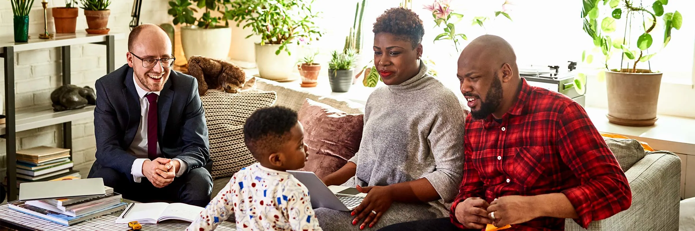  Two parents sitting on their couch with a laptop and their financial advisor. Their toddler son is looking up at the father and there are many plants in the background.
