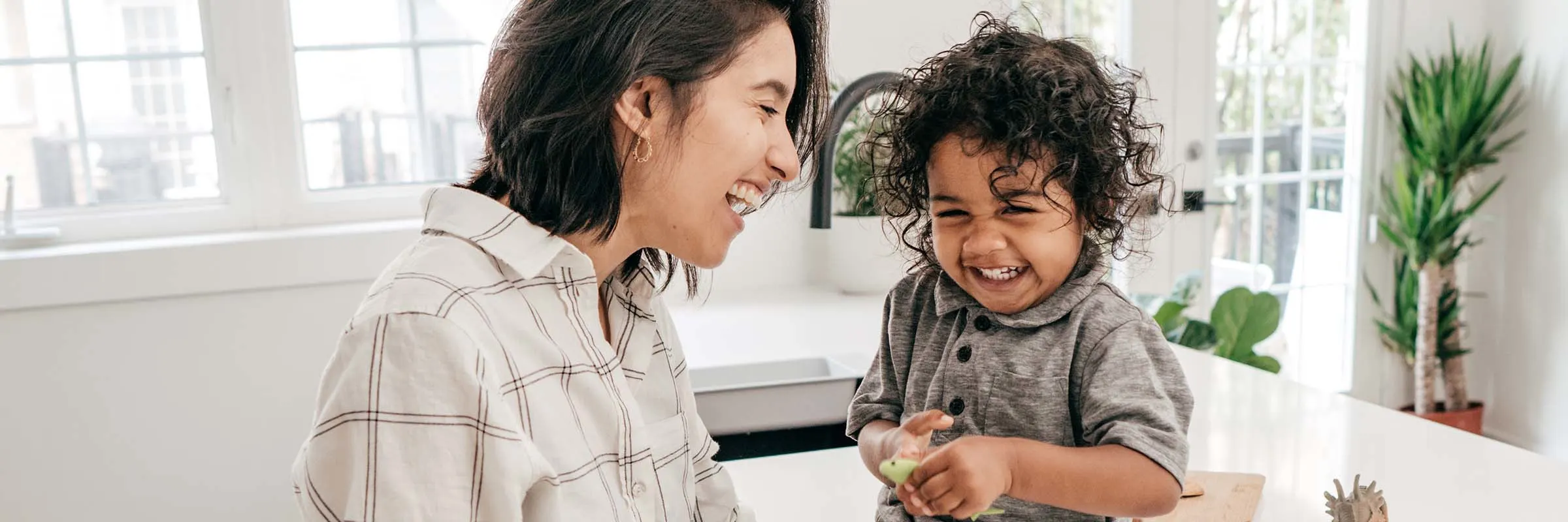  A mother and her toddler are smiling in a kitchen. The son's toys and some snacks are on the counter next to him.