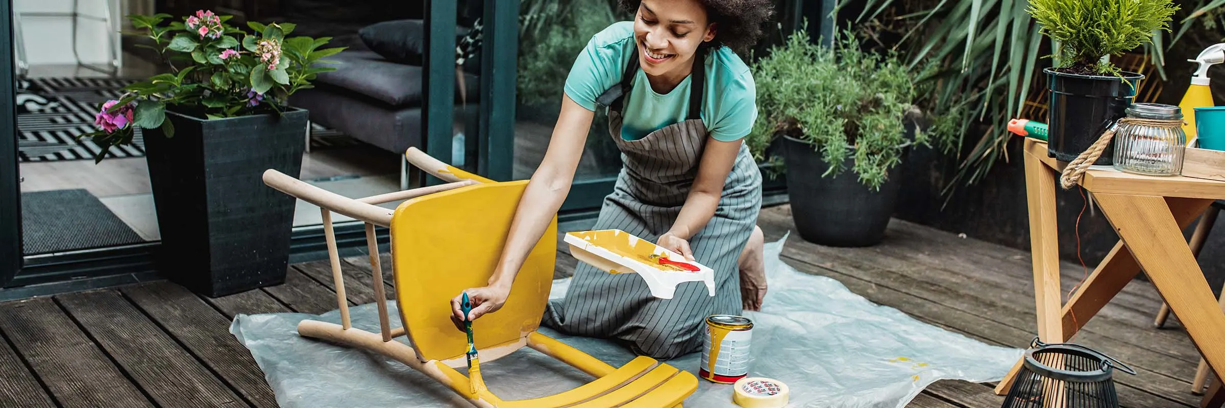 A woman smiles while she paints a wooden chair on her back patio. 