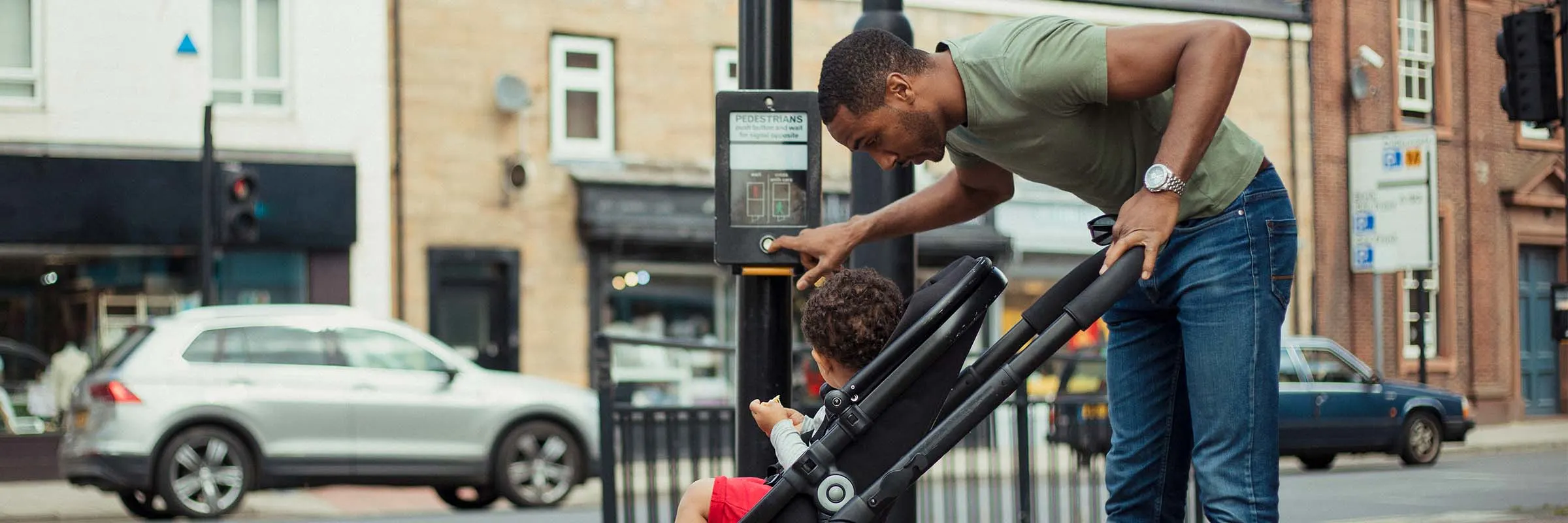 A father stands at a crosswalk and leans over to look at his son sitting in a stroller.