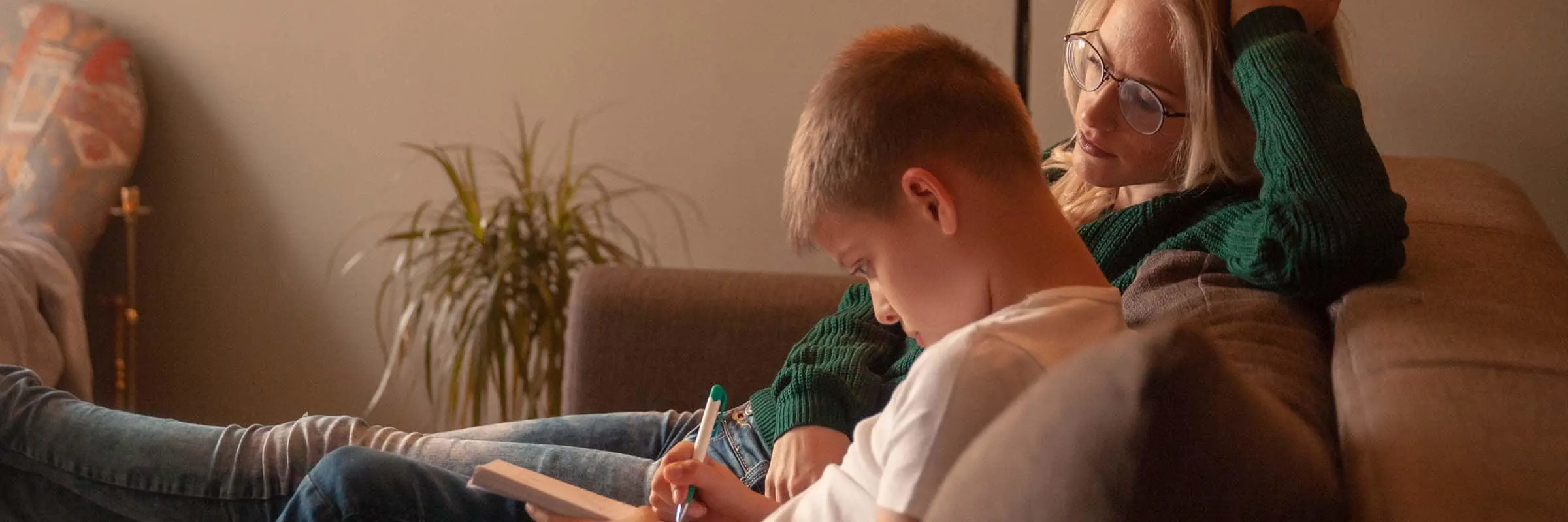 A young mother sitting on the couch with her son who&rsquo;s doing homework. 