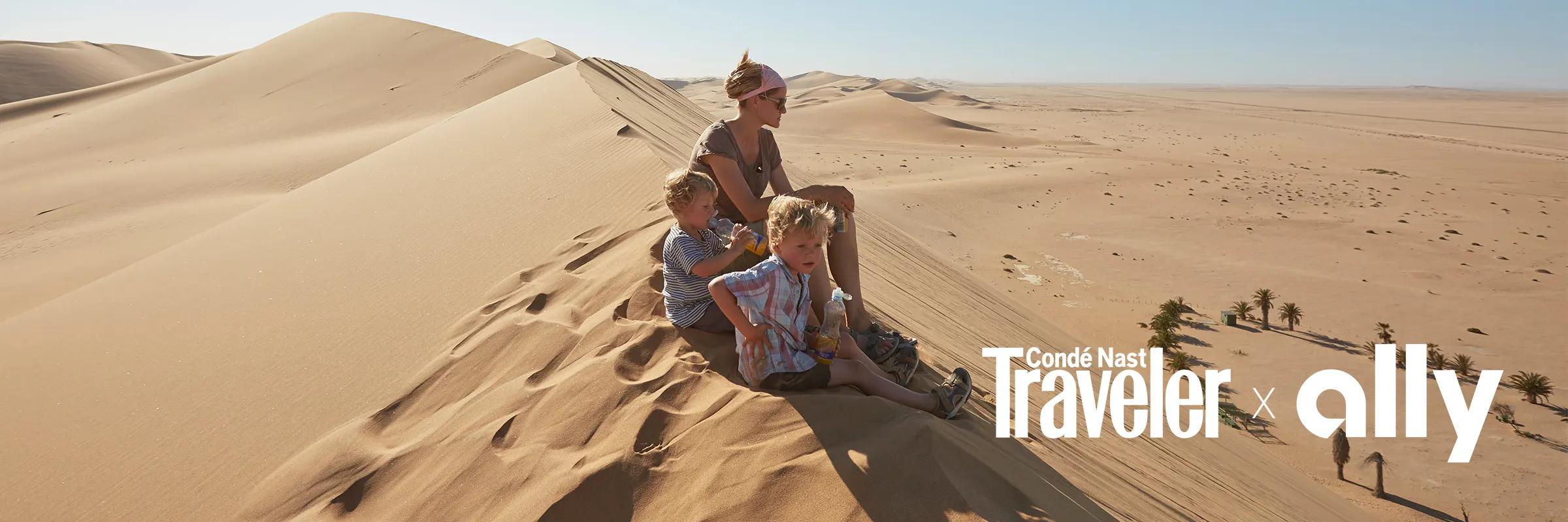 Mother and two children sit on top of a sand dune and look out over the landscape. There is a 'Conde Nast Traveler' and 'Ally' logo in the bottom right corner.