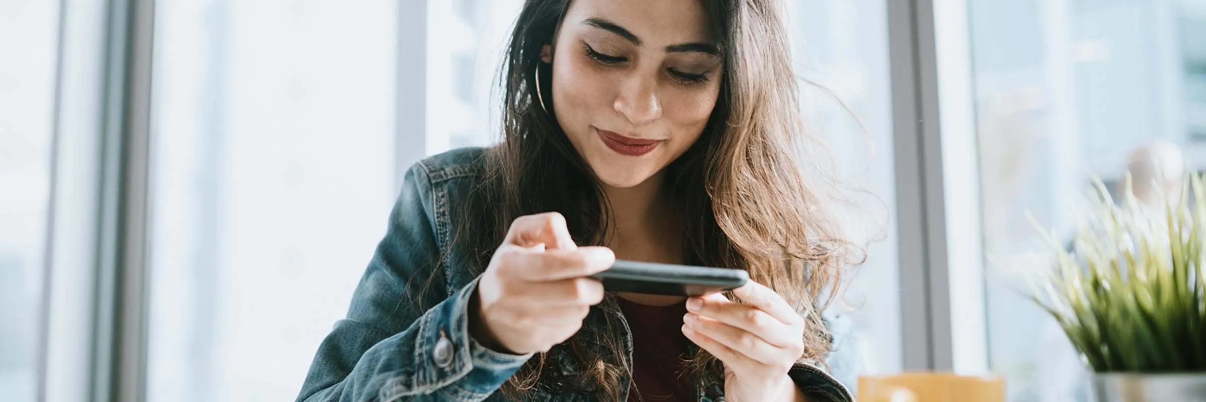 A woman uses her smartphone to electronically deposit a check. She is sitting at a table in front of large windows on a sunny day.