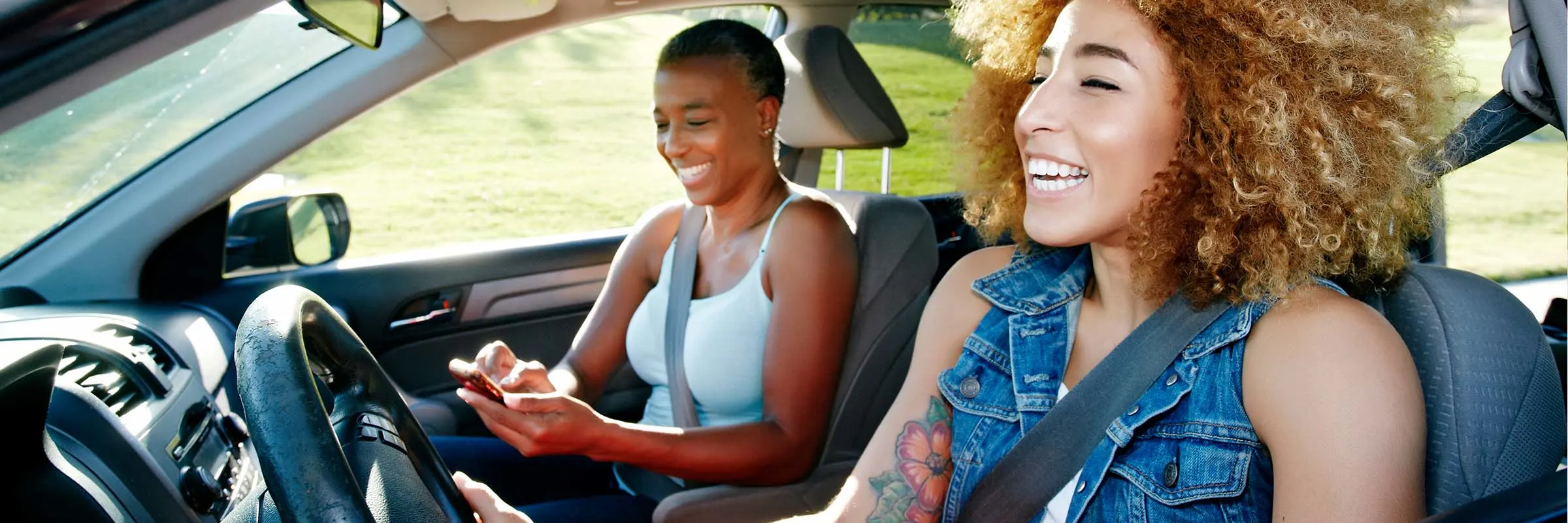 Two women in a car laughing while driving with the windows down.