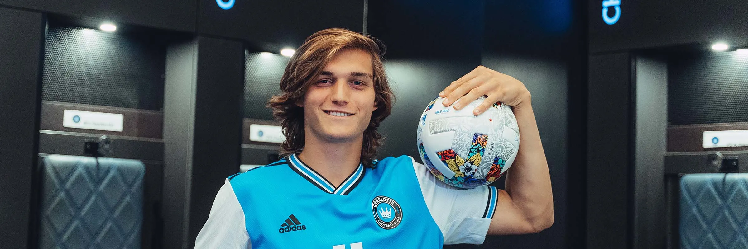 Charlotte FC midfielder, Ben Bender, poses in front of Charlotte FC logos in the team locker room while holding a soccer ball by his shoulder