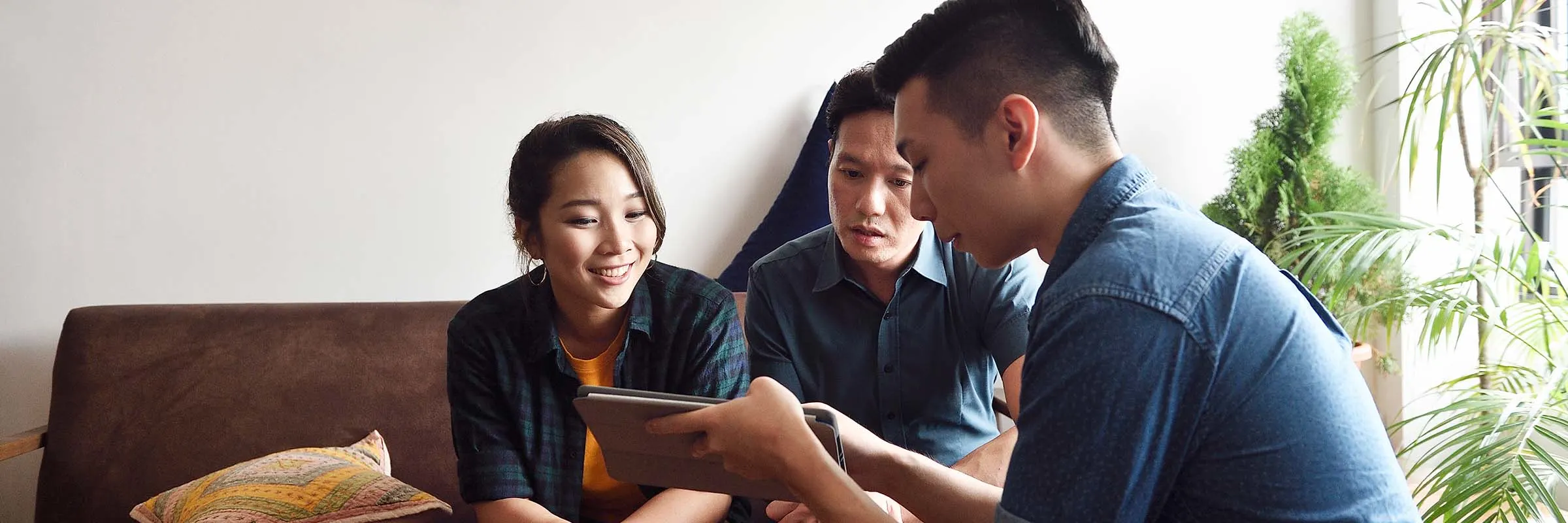 A man is showing a couple something on a tablet. All three are seated near a sunny window.