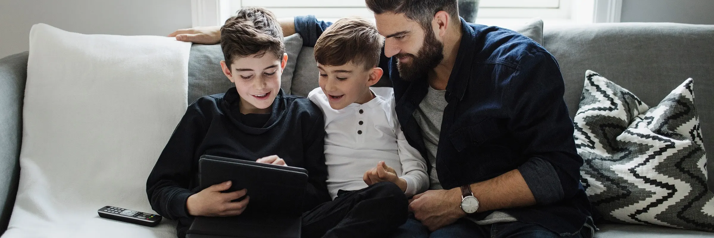 A father sitting on the couch with his two young sons while looking at a tablet.