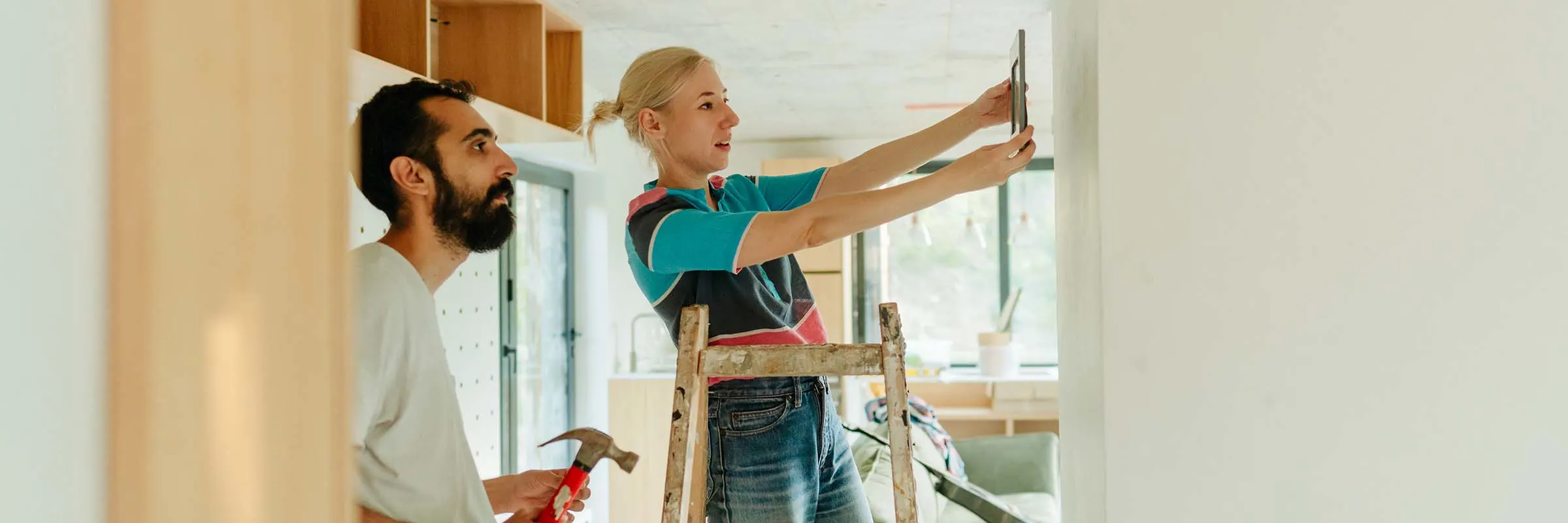 A man watches as a woman on a ladder holds a picture frame up against a wall.