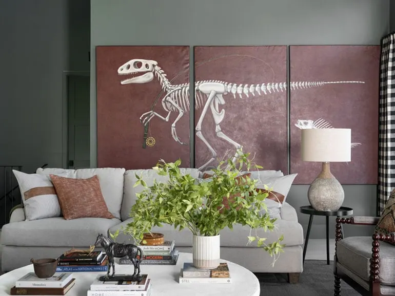 Books are stacked in small piles on a coffee table inside a living room. There is a large art piece of a dinosaur hanging on the wall in the background.