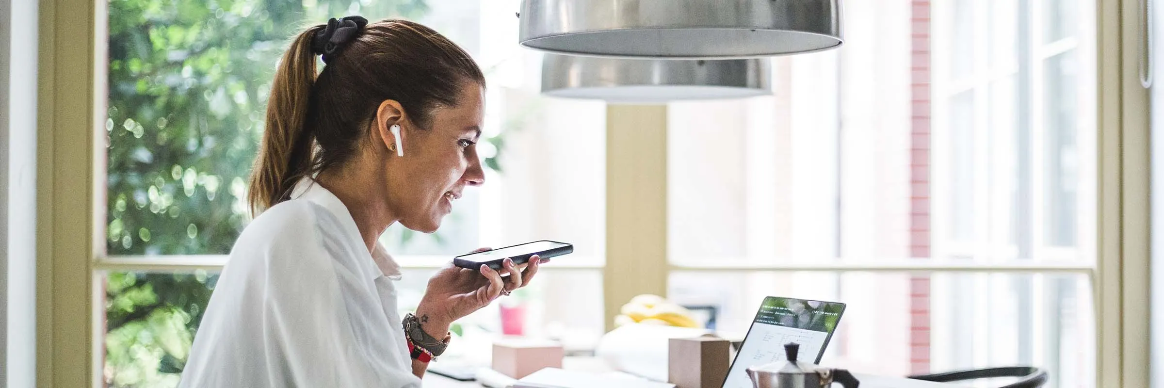 Woman with a ponytail sitting at her kitchen table on her laptop and taking a call with her wireless headphones.