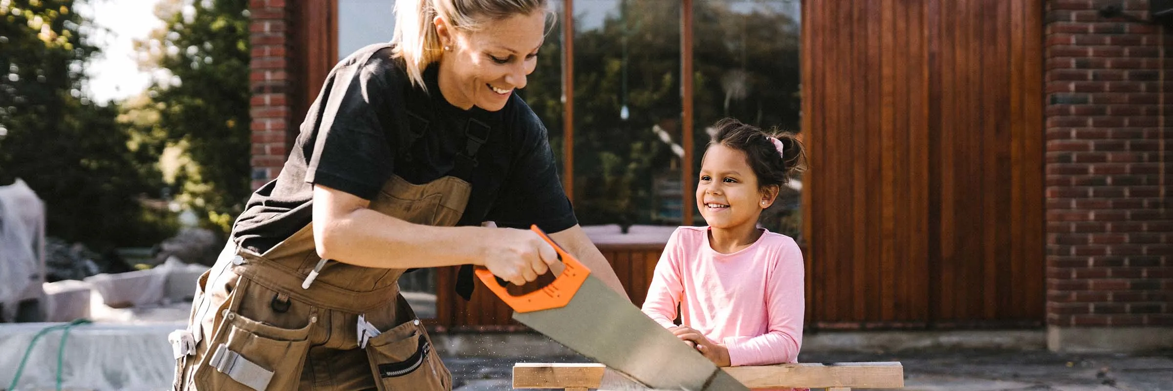 A mother is smiling while sawing a piece of wood outdoors. Her daughter is watching her.