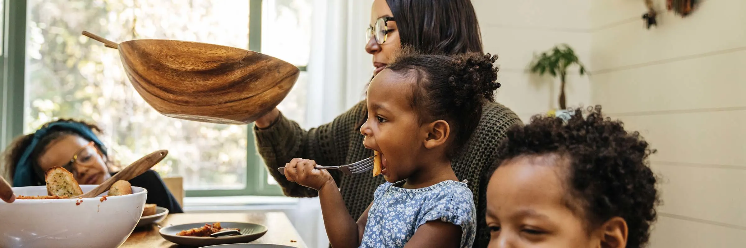 A mother is passing a salad bowl at a dining table while her three young children eat dinner. 