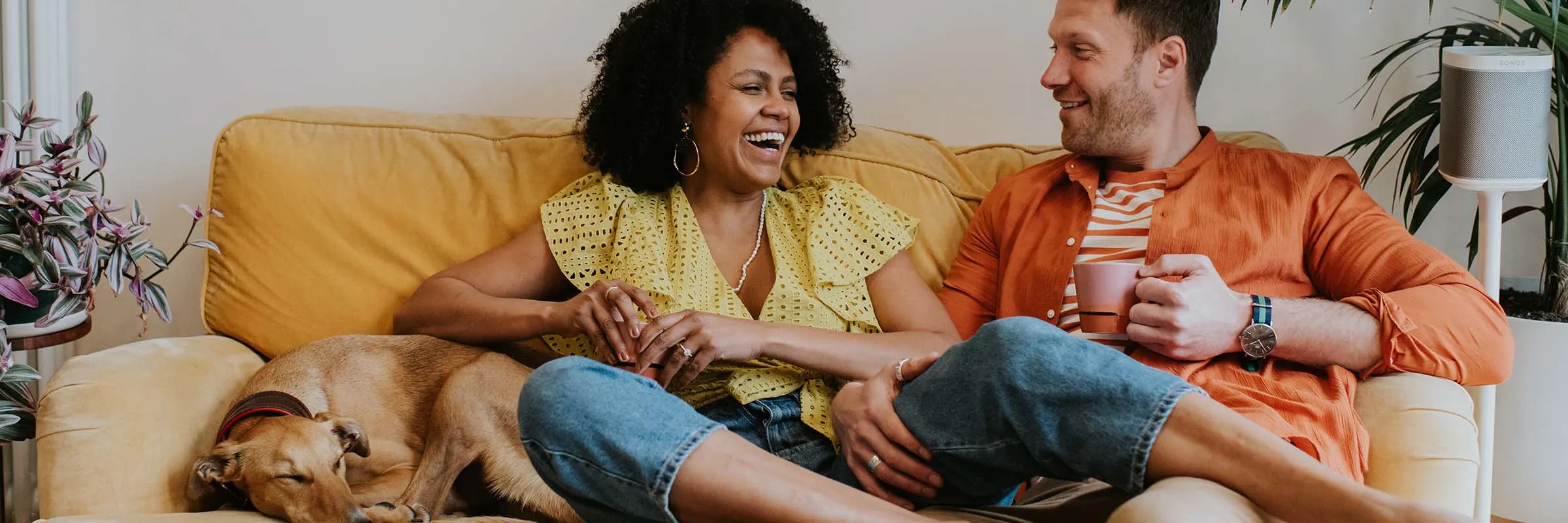 A wife, husband and dog sit on a couch together. The wife is laughing, and the husband is smiling while holding a coffee mug.