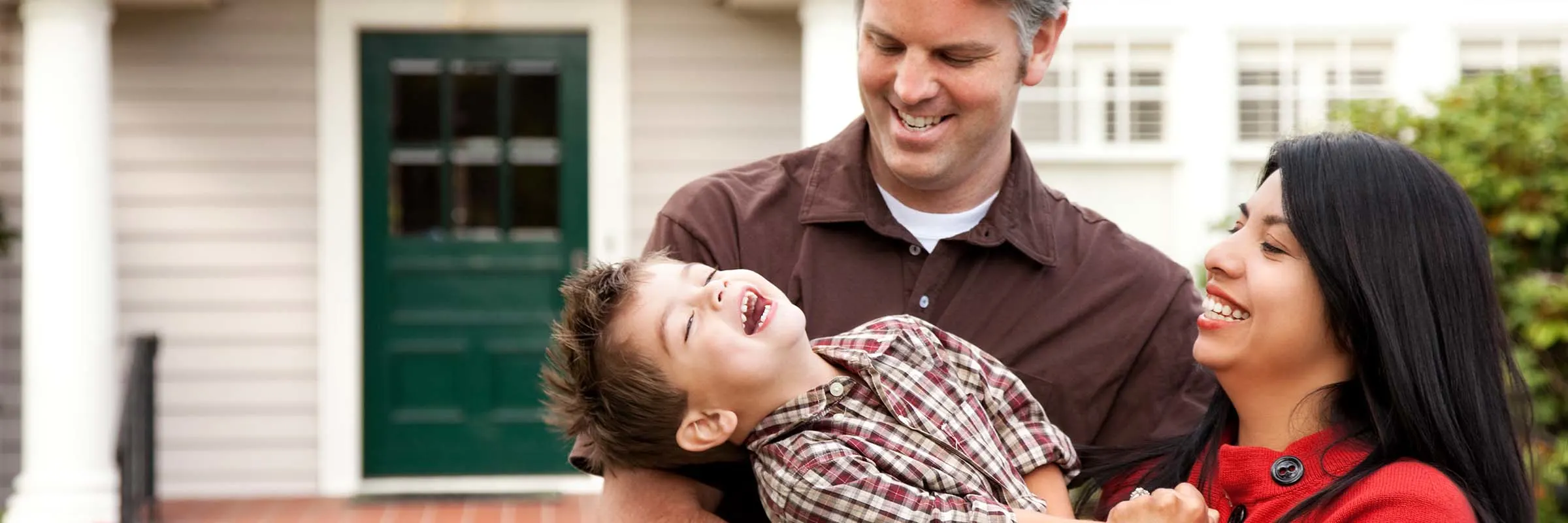 A father, mother and child laugh in front of their new home on a sunny day.