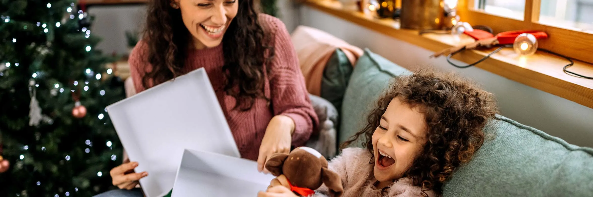 A daughter is smiling as she opens a Christmas present from her mother, who is sitting next to her on the couch.