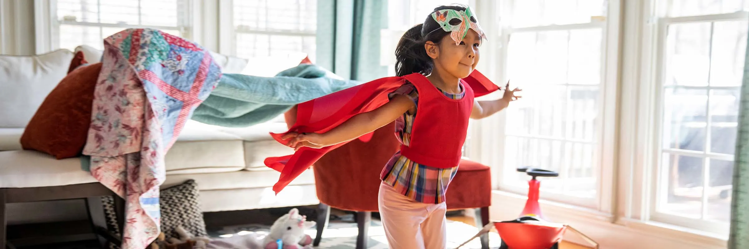 Little girl wearing a cape and a mask running through living room playing.