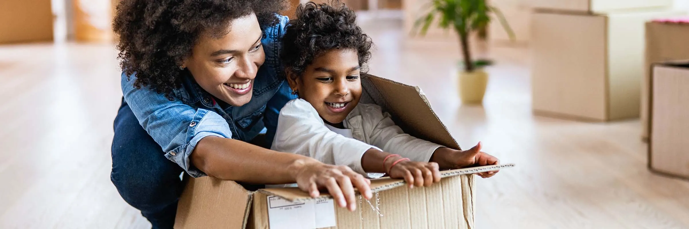 Happy mother having fun with her small girl who is inside of cardboard box at their new apartment.