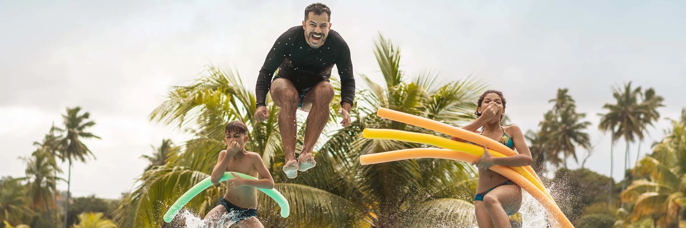 A man and two kids jumping into a pool with pool noodles