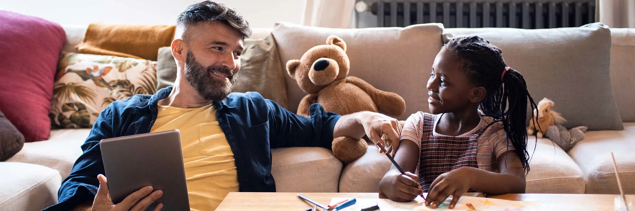 A father is looking at his daughter while she is coloring. They are both sitting on the floor in their living room.