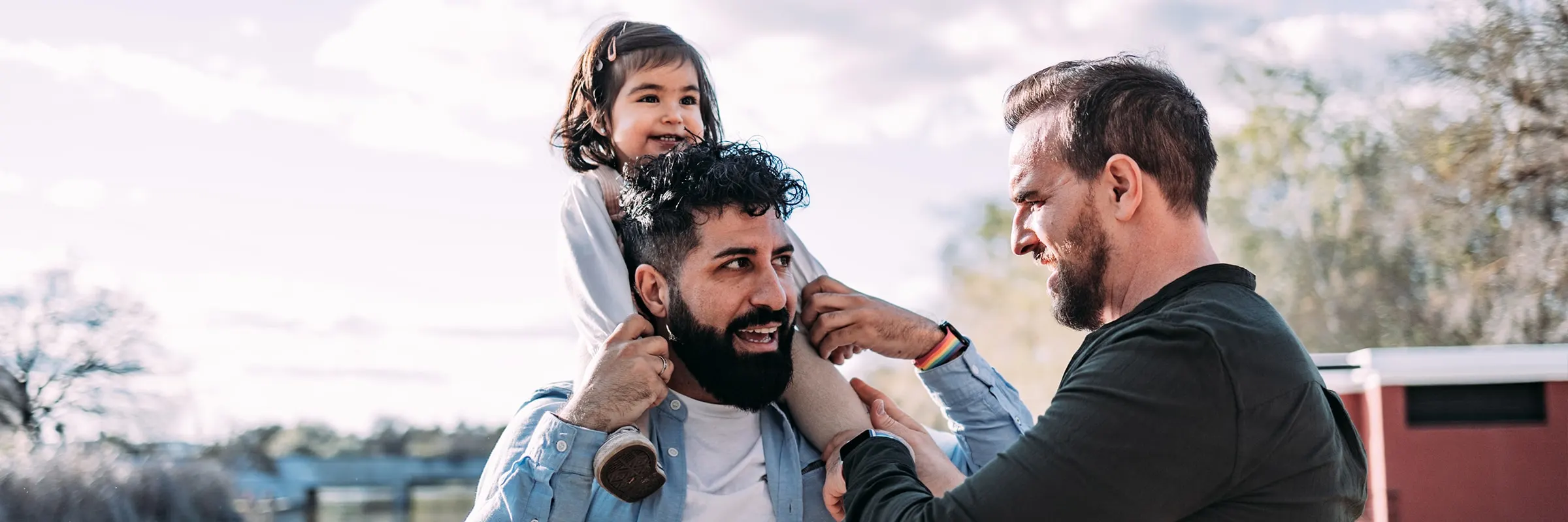  Two men with their young daughter on the shore of a lake in the park. One father holds the girl on his shoulders piggyback while the other father helps.