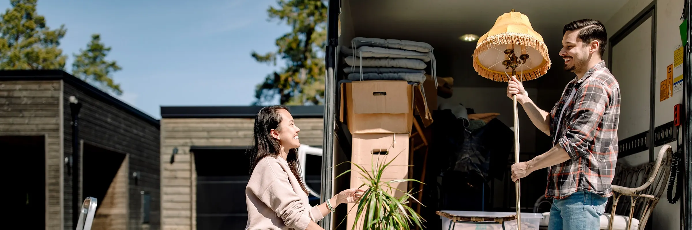 A happy couple loads cardboard boxes and furniture into a moving truck on a sunny day.