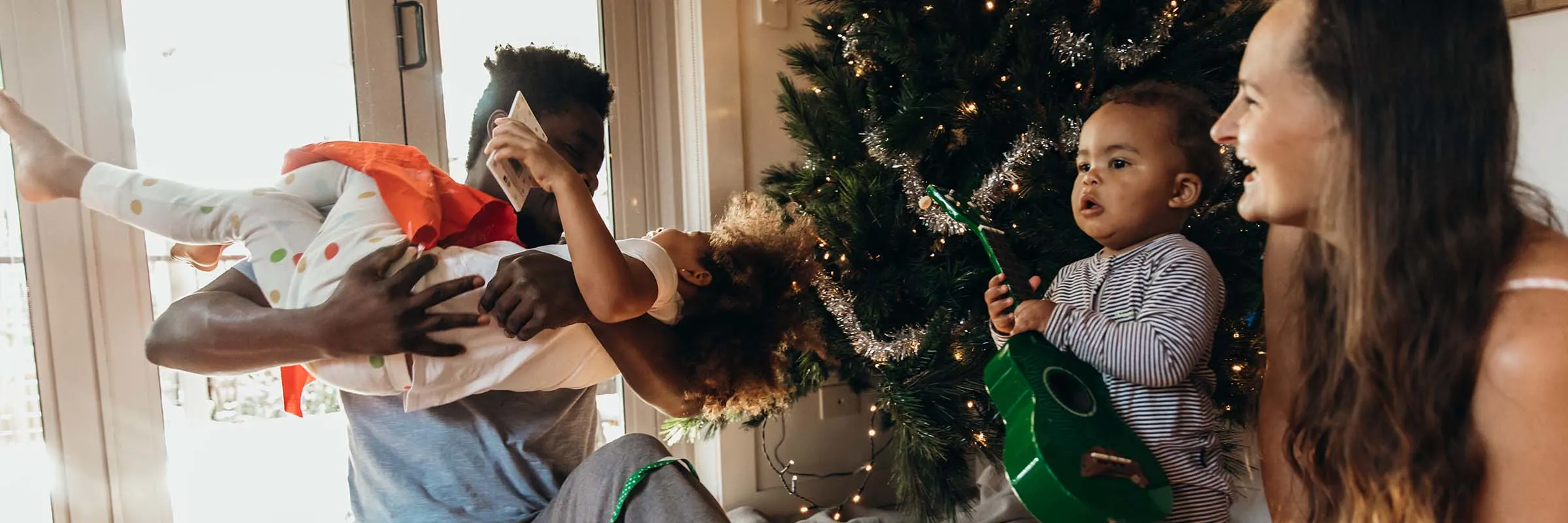 Parents and two children play with gifts under a Christmas tree.