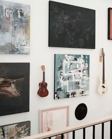 Different-sized artwork, two guitars and a hat are hung asymmetrically on a wall.