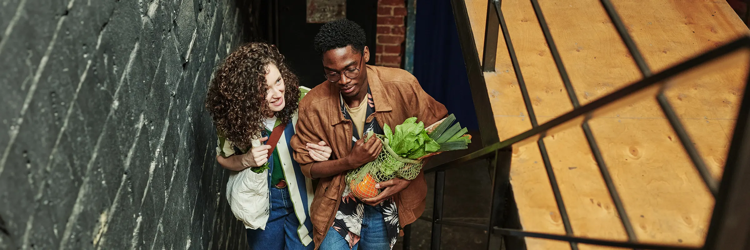 A young couple walks upstairs arm in arm, the woman smiles while the man carries a bag of groceries 