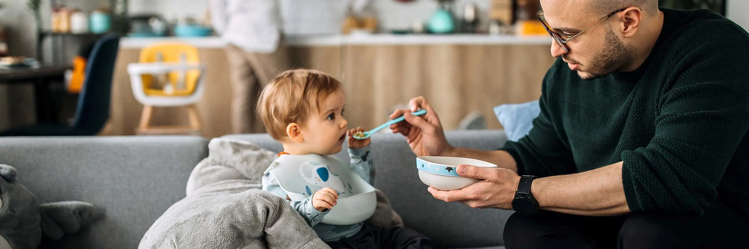 Father spoon feeding his infant son while his wife is in the background standing in the kitchen
