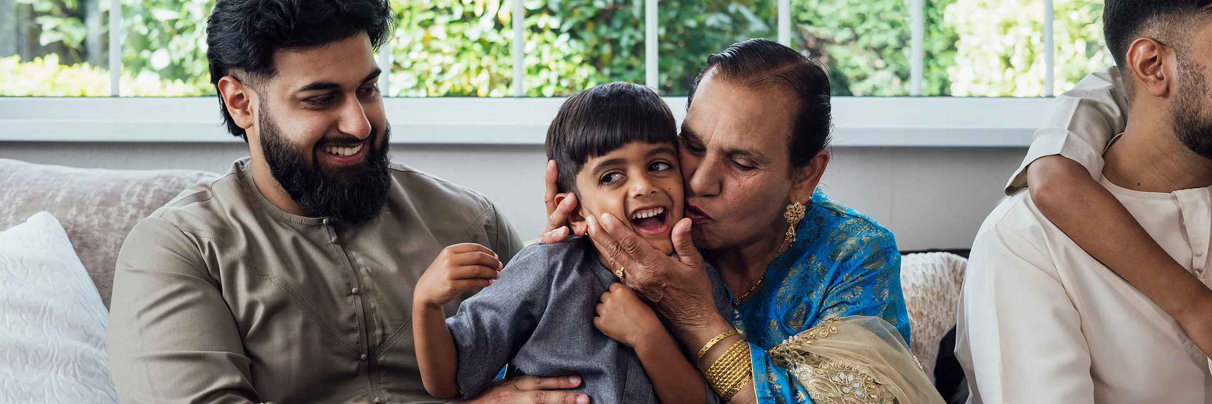 A group of family members are sitting together on a sofa, the grandmother kissing her grandchild on the cheek as they all smile. 