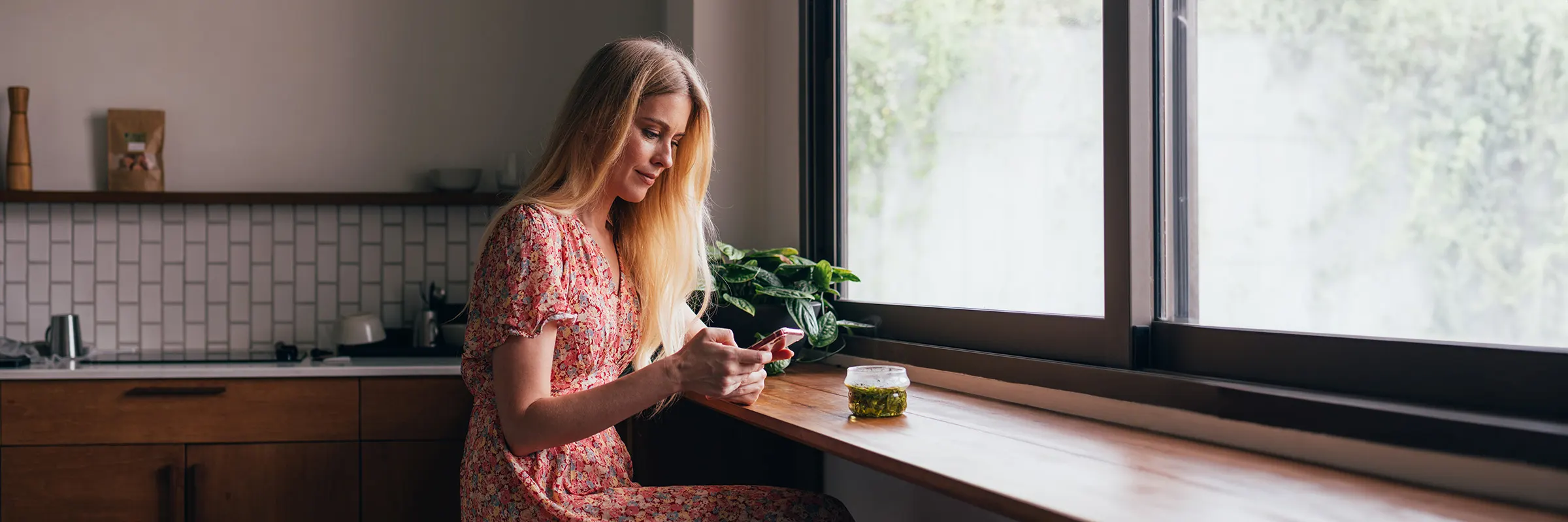  ﻿﻿﻿﻿A woman sits at a counter in the kitchen facing a large window while looking at her phone. 